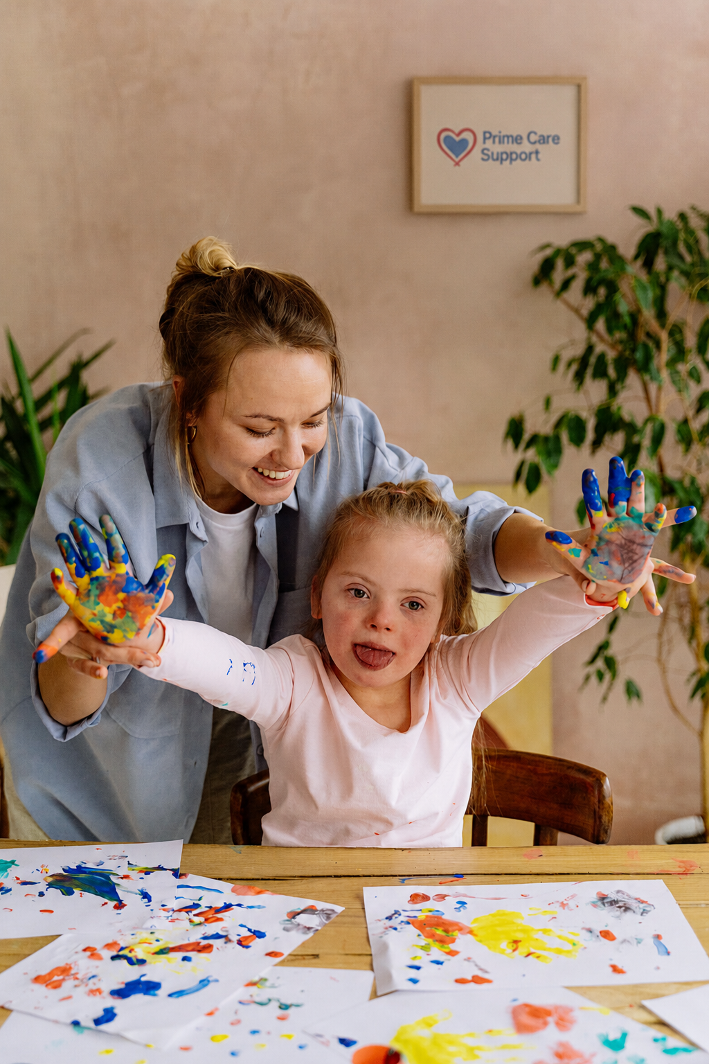 A caregiver and a young child finger-paint together at a kitchen table, both focused and laughing.